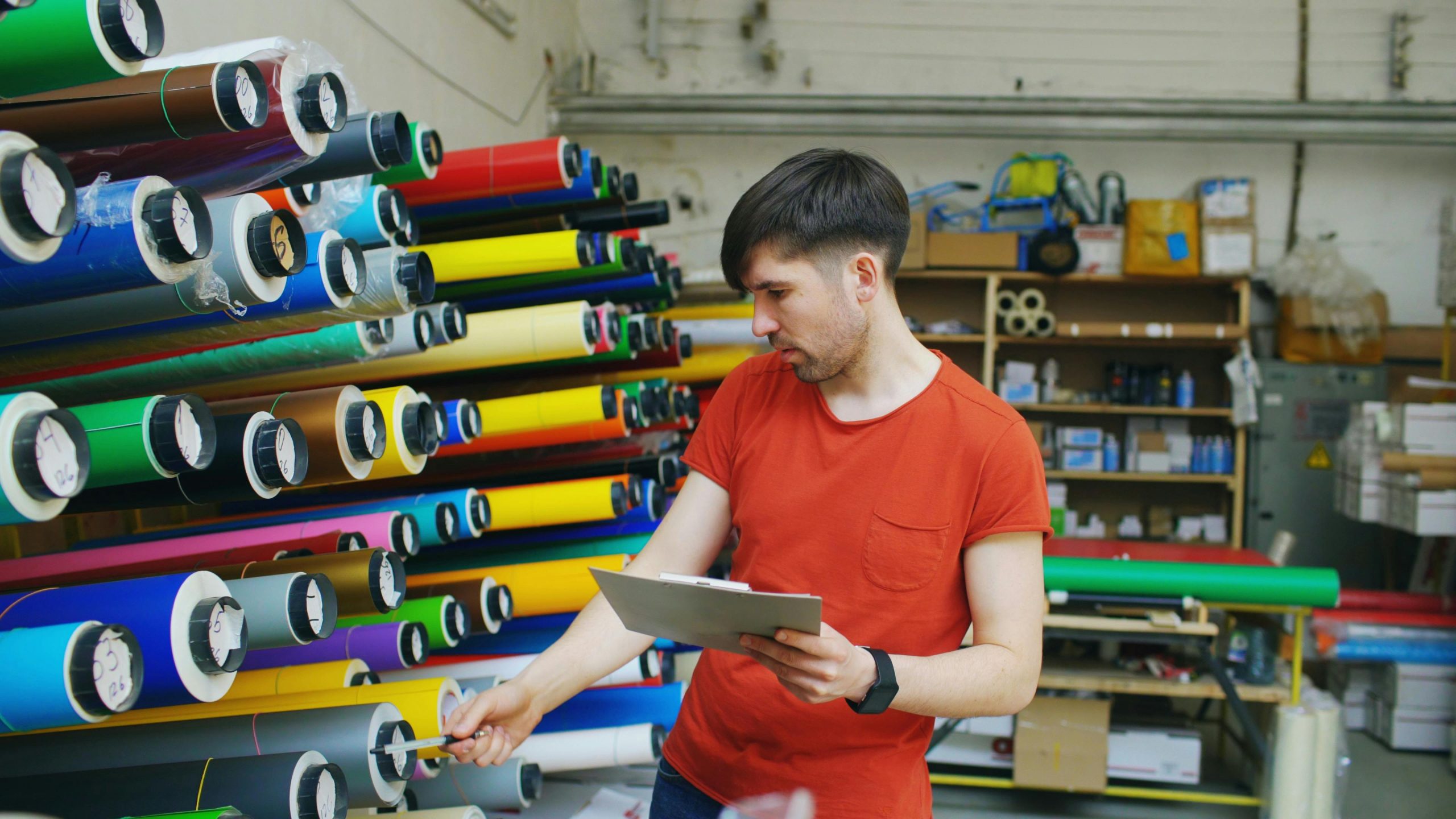 A man examining rolls of colorful vinyl in an industrial warehouse setting.